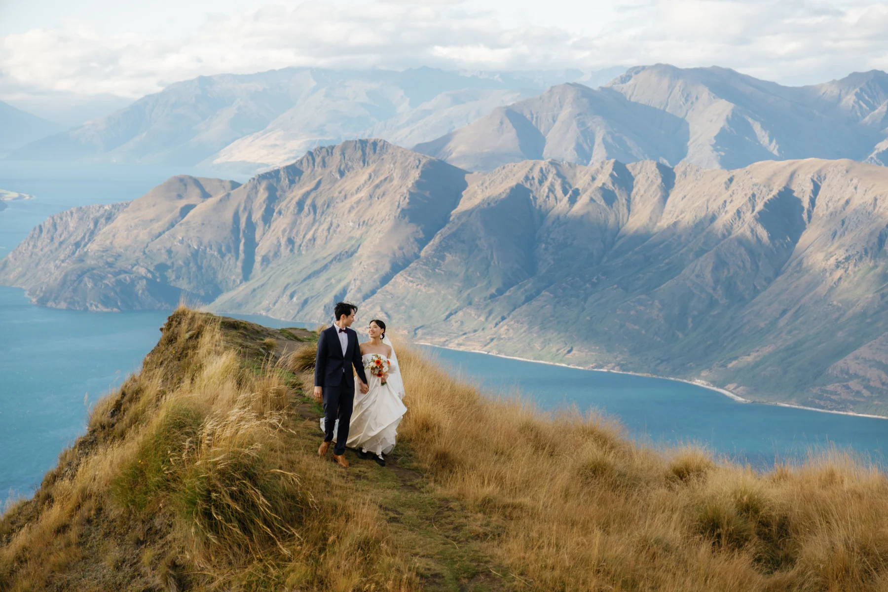 The bride and groom walking hand in hand along a ridge above the lake