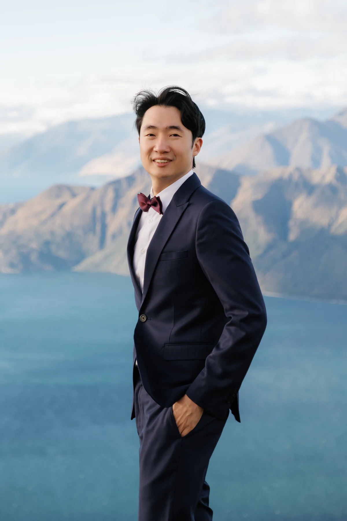 The groom in a dark suit standing before a lake and mountains