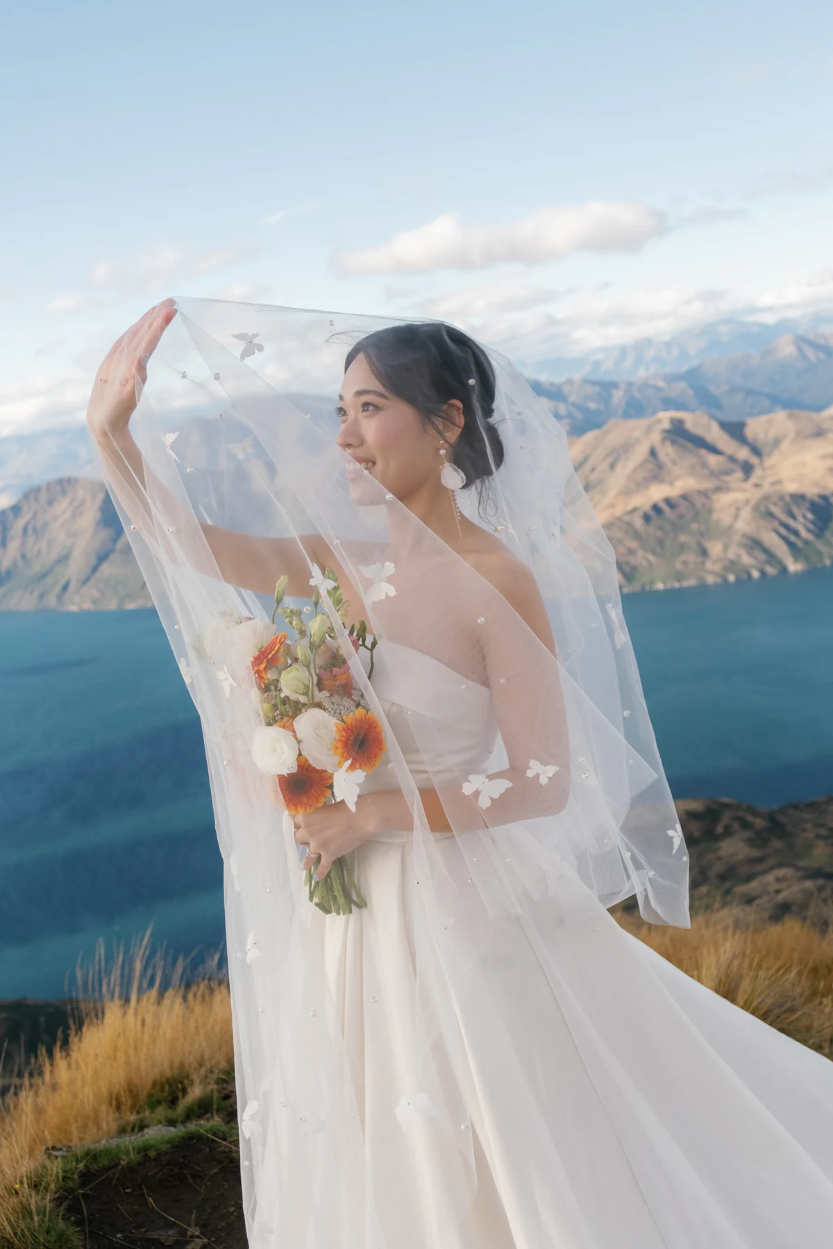 The bride holding a bouquet and lifting her veil before a lake and mountains